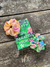 Load image into Gallery viewer, two colorful scrunchie with floral pattern, placed on a wooden surface, with a book and a flower in the background.