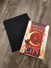Load image into Gallery viewer, A black book sleeve with a minimalist design, displayed next to a book with a vibrant red and gold cover design.