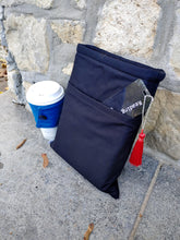 Load image into Gallery viewer, A black book sleeve with a front pocket, made of cotton fabric, displayed beside a cup and a stone surface.