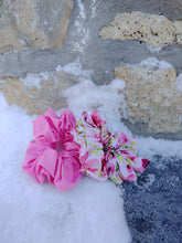Load image into Gallery viewer, A blush pink scrunchie beside a floral pattern scrunchie, resting on a textured surface with a stone wall in the background.