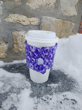 Load image into Gallery viewer, A purple floral print drink cozie wrapped around a white coffee cup, placed on a stone surface with a brick wall in the background.