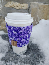 Load image into Gallery viewer, A purple floral print drink cozie wrapped around a white coffee cup, placed on a stone surface with a brick wall in the background.